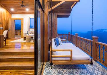 Wooden swing bed hangs on a chain, overlooking a misty mountain view. It's on a tiled balcony attached to a cozy room with wooden floors and warm lighting.