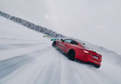 A red sports car drifts on a snow-covered track, closely followed by two others. Snow swirls around, and trees line the horizon. Text on the car reads 