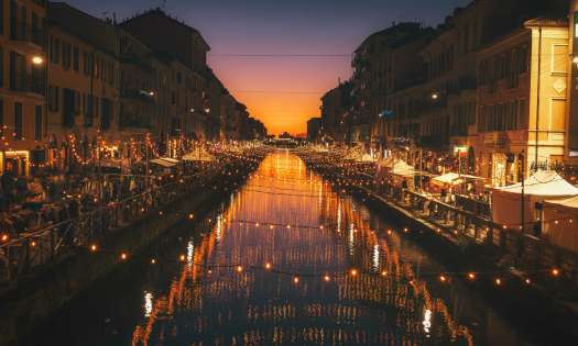 String lights over a river with street shops on either side during sunset.