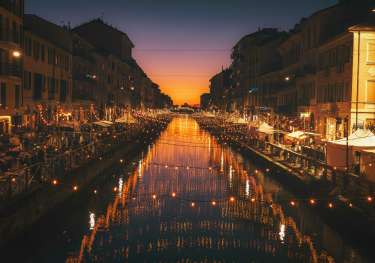 String lights over a river with street shops on either side during sunset.