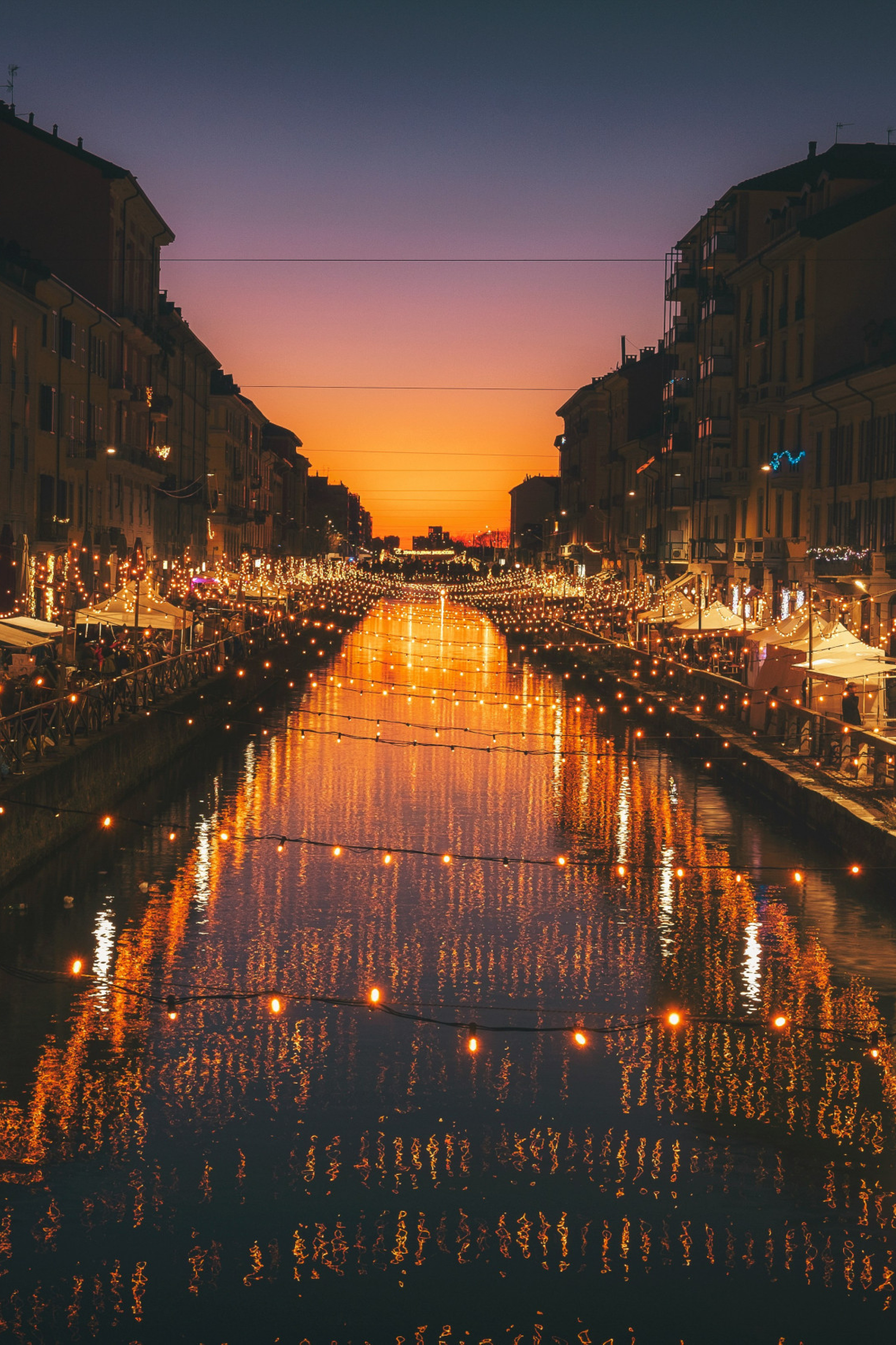 string lights over a river with street shops on either side during sunset