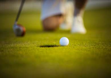 Golf ball on the outside of a hole with a man kneeling behind, holding a golf club.