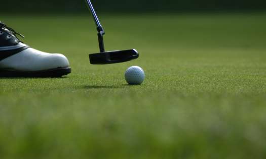 Golf ball and a small golf club with a shoe to the left on grass.