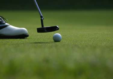 Golf ball and a small golf club with a shoe to the left on grass.