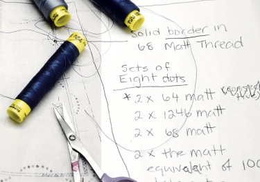 Three thread spools rest on a sketch and notes about making a tablecloth. Nearby, scissors lie on paper with text describing dimensions and thread details for different sections.