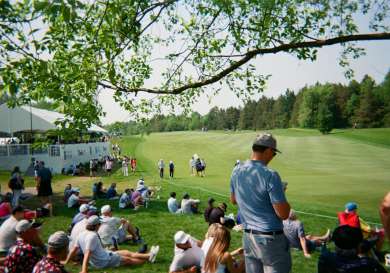 Crowd of people sitting/standing and looking out at a golfing green.