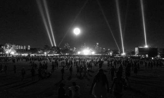 Black and white image of a park at night with strobe lights and a bright moon above a stage.