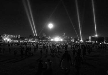 Black and white image of a park at night with strobe lights and a bright moon above a stage.