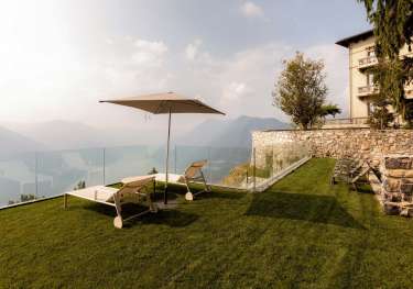 Sun loungers rest under a beach umbrella on a grassy terrace, overlooking distant mountains. The area is bordered by a glass railing and stone wall, adjacent to a building.