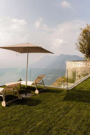 Sun loungers rest under a beach umbrella on a grassy terrace, overlooking distant mountains. The area is bordered by a glass railing and stone wall, adjacent to a building.