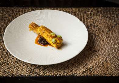 Grilled corn coated with spices rests on a white plate, garnished with chopped green onions; the plate is set on a woven, neutral-toned placemat.