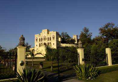 A large, elegant castle stands amidst lush gardens, bordered by decorative wrought-iron gates and flanked by trees, under a clear blue sky.