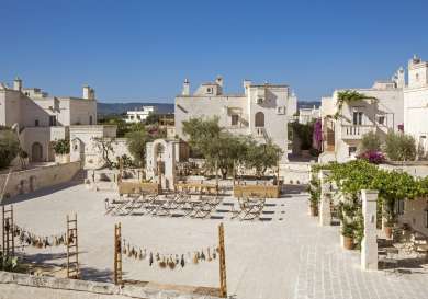 A rustic courtyard features wooden tables and chairs arranged under olive trees, surrounded by white stone buildings adorned with vines and flowers, under a clear blue sky.