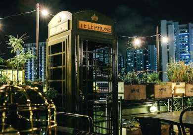 A vintage green telephone booth stands unused on a rooftop garden at night, surrounded by plants and wooden crates, with city skyscrapers illuminated in the background.