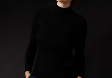 A person smiles widely, wearing a black turtleneck and red plaid pants, hands in pockets, set against a plain dark background.