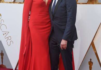 A woman in a red gown kisses a smiling man in a suit on the cheek.