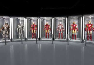 Seven Iron Man suits stand displayed in illuminated glass cases, each uniquely designed, within a modern, sleek exhibition hall.