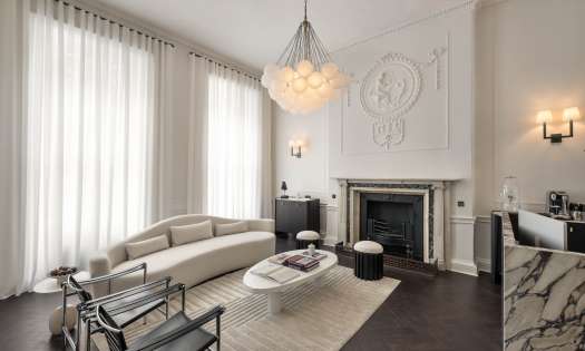 A modern living room features a curved sofa facing a fireplace. Above hangs a round chandelier. The room is elegantly decorated with tall curtains and stylish furniture.