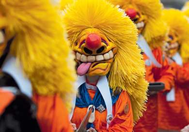 A person wears a vibrant, exaggerated clown mask with yellow hair and orange attire.