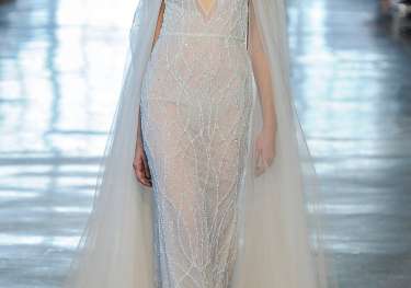 A model walks in a sheer, embellished gown with a flowing cape, under soft lighting, on a runway lined with seated spectators.