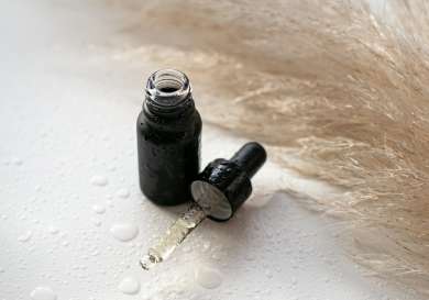 A black dropper bottle rests on a wet surface, surrounded by light beige pampas grass, with droplets of liquid scattered around.