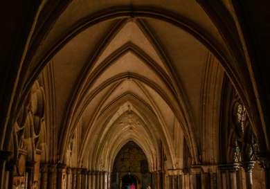 Arched stone hallway with ribbed vaults casts long shadows; people sit and walk along benches beneath stained glass windows in a historic, dimly lit architectural setting.