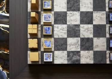 Chess pieces with abstract geometric designs are arranged on a checkered board, positioned on a wooden table with a decorative, marbled edge.