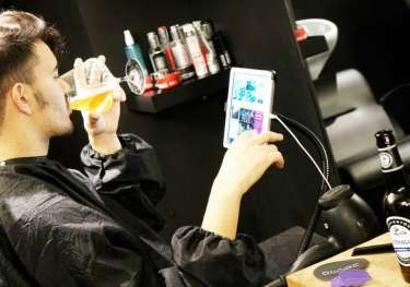 A person in a barber's chair drinks beer while viewing a tablet.