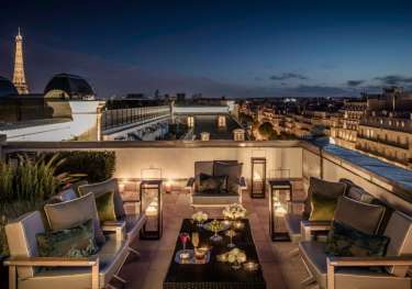 Elegant rooftop patio with chairs and tables adorned with drinks and flowers, softly lit by lanterns.