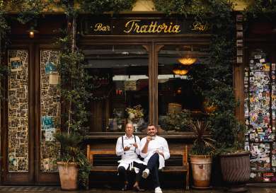 Two people sipping coffee on bench outside restaurant .