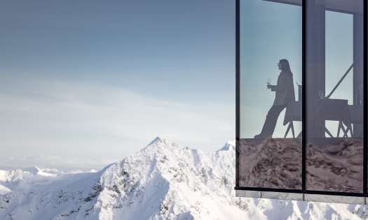 Photo of person drinking wine in restaurant with glass walls looking over snowy mountains.