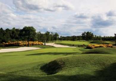 Rolling green hills stretch across a golf course with sand bunkers, surrounded by trees and flowering shrubs under a partly cloudy sky.