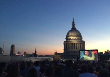 A crowd watches an outdoor movie on a large screen at dusk, with a historic domed building and city skyline in the background under a clear evening sky.