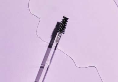 A clear mascara wand lies diagonally on a pastel purple surface, next to an amorphous, shiny liquid spill, casting soft shadows.