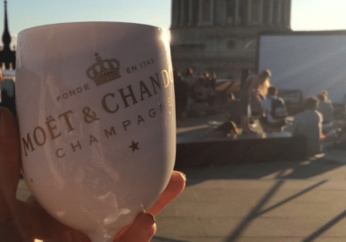 A white Moët & Chandon champagne glass is held up against a backdrop of people relaxing on a rooftop under a clear sky, with a domed building in the background.