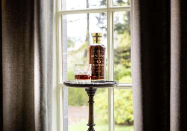 Bottle of whisky and whisky glass styled next to window .