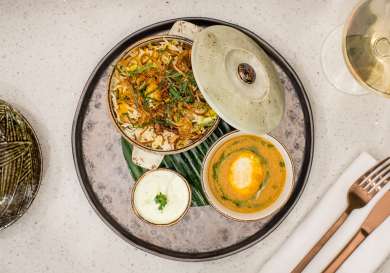 A lidded bowl of biryani and a small bowl of creamy curry rest on a textured round plate, accompanied by yogurt sauce.