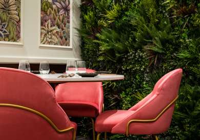 Pink chairs surround a round table set with glasses and cutlery, positioned against a lush green plant wall, with botanical art hung on the adjacent wall.