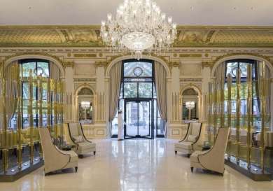 A grand chandelier hangs in an ornate hotel lobby with high ceilings, elegant seating, and floor-to-ceiling windows that offer a view of tree-lined streets.