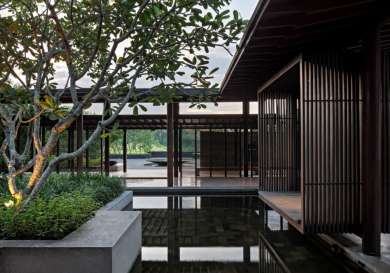 A tree stands beside a reflective pool, surrounded by wooden structures with vertical slats, set in a tranquil, open-air architectural space with lush greenery in the background.