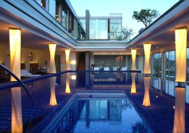 A luxurious indoor pool reflects illuminated columns surrounded by modern architecture, with large glass windows and seating areas on either side, providing a serene and elegant ambiance.