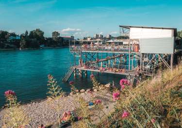 A riverside structure on stilts hosts people sunbathing and socializing.