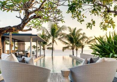 Two wicker chairs face an infinity pool reflecting palm trees, with a covered outdoor seating area nearby.