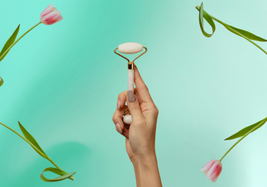 Person holding face roller against green background framed by pink tulips.