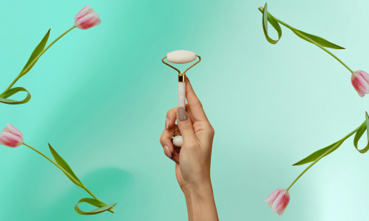 Person holding face roller against green background framed by pink tulips.