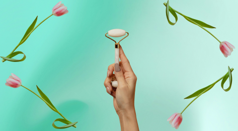 Person holding face roller against green background framed by pink tulips.