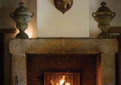 A fireplace with a burning fire is centered between two ornate urns; a mounted deer head hangs above on a stone wall.