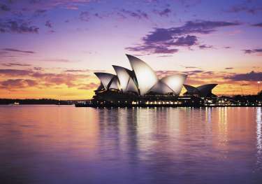 Iconic white sails of the Sydney Opera House are illuminated against a colorful sunset, reflecting on the calm harbor waters, creating a serene and picturesque scene.