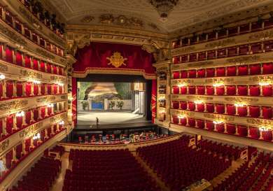 An opulent theater stage featuring a scenic backdrop is set for a performance, surrounded by ornate balconies with red velvet seats and golden decorations, viewed from the audience's perspective.