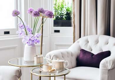 Cream chairs inside hotel with two round tables with teapot and purple flowers.